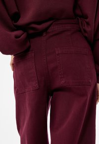 Burgundin denim-housut, joissa on kaksi takataskua, suora lahkeensuukuvio ja tekstuuri, jossa on näkyviä ompelusaumoja.