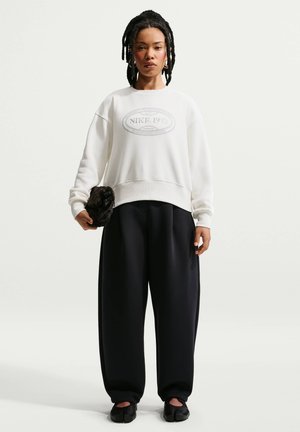 Person iført en hvid Nike 1972 sweatshirt, løse sorte bukser, sorte sko, holder en lille sort tekstureret taske, stående mod en ensfarvet baggrund.