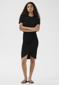 T-shirt nero a maniche corte abbinato a una gonna avvolgente nera arricciata, completata con uno spacco e indossata con infradito nere.