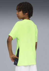 Bambino con capelli corti intrecciati che indossa una maglietta sportiva verde lime con pannelli laterali neri, in piedi di lato con una mano dietro la schiena.