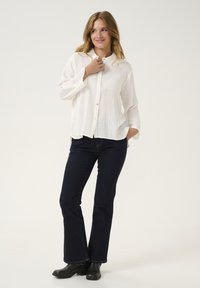 White button-up shirt with a crinkled texture, paired with dark blue jeans and black ankle boots, showcasing a relaxed fit and side slits.