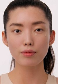 Modèle avec des cheveux noirs raides, peau claire, fond de teint léger appliqué sous les yeux et sur les joues, couleur de lèvres neutre, portant un haut beige.