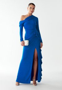 Pikk sinine kleit ühele õlale, ruffle-accentidega ja reie kõrgune lõhik. Valmistatud siledast kangast, koos valge käekoti ja hõbedaste kontsakingadega.