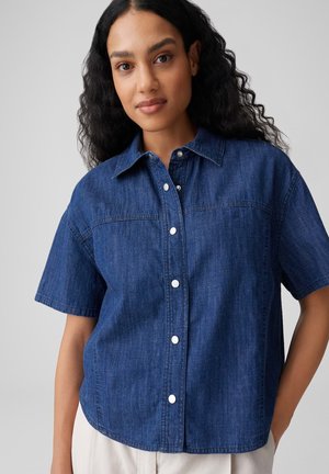 Frau mit langen lockigen Haaren, die ein kurzärmliges, blaues Denim-Hemd mit Knöpfen trägt, steht vor einem schlichten hellgrauen Hintergrund.