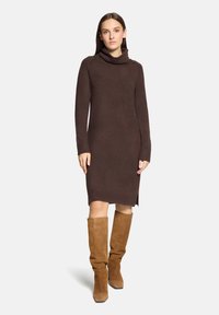Femme portant une robe-pull marron à col roulé et manches longues, ainsi que des bottes hautes en daim beige, debout devant un fond clair uni.