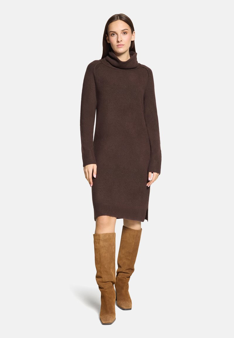 Femme portant une robe-pull marron à col roulé et manches longues, ainsi que des bottes hautes en daim beige, debout devant un fond clair uni.