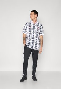 White and black patterned short-sleeve shirt with hexagonal designs, paired with black pants and shoes, featuring minimal branding accents.