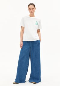 Jeune femme portant un T-shirt blanc avec du texte et un jean bleu à jambes larges, se tenant avec les mains détendues, expression neutre, fond blanc uni.
