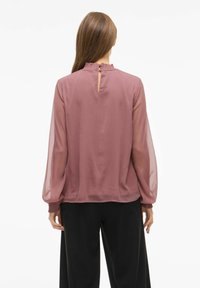 Blusa semitranslúcida de color rosa, con largas mangas abullonadas, cuello con volantes y detalle de ojal en la parte posterior, combinada con pantalones negros.