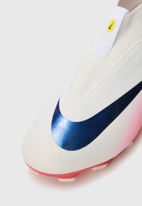 Crampon de football blanche avec un swoosh Nike bleu marine brillant. Présente un dessus texturé, une semelle en dégradé de rose et une étiquette jaune sur la languette.