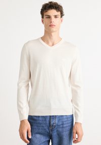 Maglione crema a v in fine maglia, con maniche lunghe e orlo a coste, indossato con jeans blu. Logo sottile sul petto.