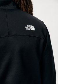 Sudadera negra con una textura suave y un logo blanco en la parte superior de la espalda. Presenta un cuello alto y un diseño contemporáneo.