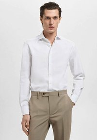 Camisa de vestir blanca de manga larga con cuello abotonado, combinada con pantalones beige. La camisa presenta una textura suave y un diseño ceñido.