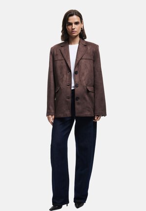 Braune Wildlederblazer mit lockerer Passform, großen Knöpfen, zwei vorderen Taschen und einem einreihigen Design. Kombiniert mit dunklen Jeanshosen.