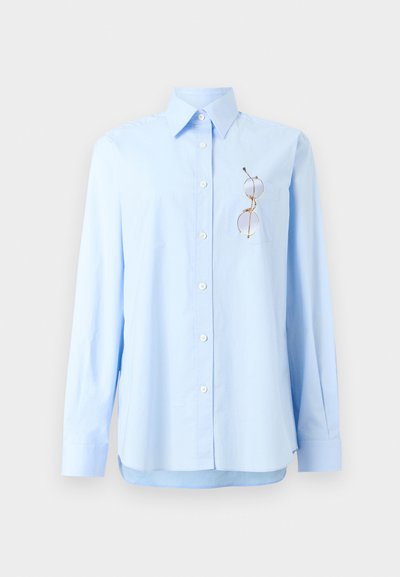 Chemise bleu clair à manches longues avec col et poche contenant une paire de lunettes à monture dorée, présentée sur un fond blanc.