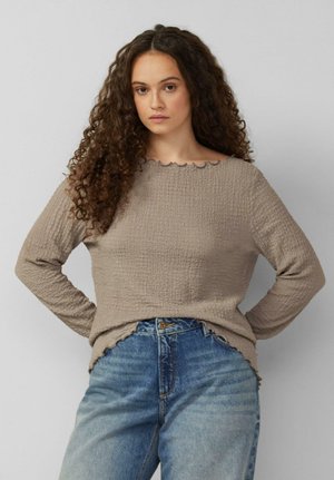 Kobieta o długich, kręconych włosach, ubrana w beżową, teksturowaną bluzkę z długim rękawem i niebieskie jeansy z wysokim stanem, stoi z rękami na biodrach.