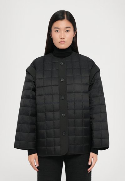 Polstret sort jakke med quiltet tekstur, oversized form, rund halsudskæring og knaplukning foran. Båret over en sort turtleneck.