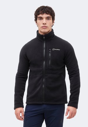 Man draagt een zwarte Berghaus fleecejas met ritssluiting, staand tegen een effen lichte achtergrond, naar voren kijkend.