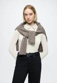 Hnedý vlněný šál prehodený cez svetlo béžovú blúzku, s koncami zviazanými dokopy. Oblečenie je doplnené čiernymi nohavicami a čiernym opaskom.