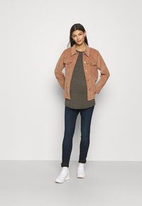 Jacket de corduroy castanho claro com dois bolsos frontais, usado por cima de uma camisa riscada escura, combinado com jeans escuros e ténis brancos.