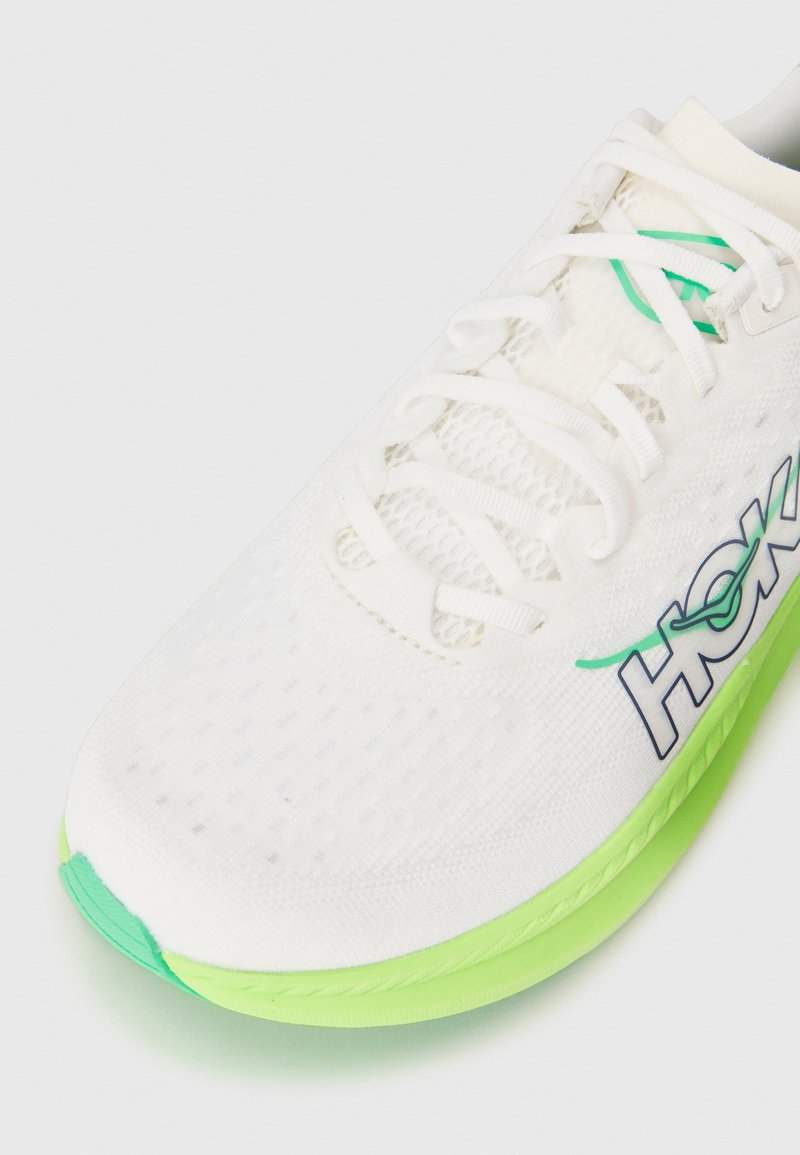 Vita mesh löparsko med gröna accenter och en texturerad sula, med snörning och en framträdande logotyp på sidan.