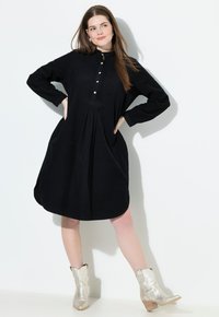 Abito lungo nero a maniche lunghe con scollatura alta, chiusura con bottoni, gonna plissettata e tasche laterali. Presenta una texture morbida e una vestibilità comoda.