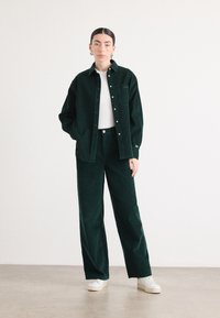 Dunkelgrünes Cordhemd mit Knöpfen und passenden weit geschnittenen Hosen, kombiniert mit weißen Sneakers, mit einer Fronttasche. Glatte Textur.