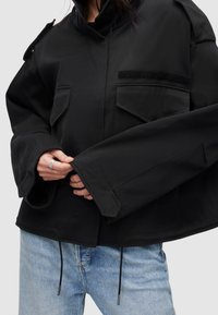 Zwarte, oversized jas met een hoge kraag, twee voorzakken en een gestructureerd patch. De stof is glad en heeft verstelbare trekkoorden aan de zoom.