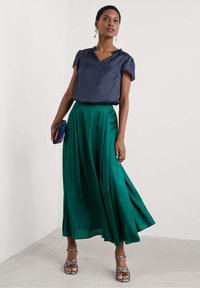 Navy Satin-Oberteil mit kurzen Ärmeln; grüne fließende Maxiröcke; silberne Riemchensandalen; hält eine kleine Clutch mit blauen und roten Akzenten.