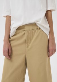 Personne portant un pantalon beige ample et une chemise blanche à manches courtes, les mains reposant légèrement sur les côtés.
