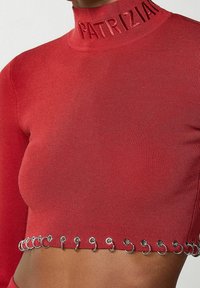 Červený žebrovaný crop top s vysokým límcem se zlaceným nápisem "PATRIZIA" a dekorativním stříbrným kovovým detailem podél lemu.
