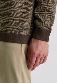 Braun gemusterter Strickpullover mit einem strukturierten gewebten Design; hat einen dunkelbraunen gerippten Saum und ein kleines Logo-Tag an der Seite.