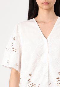 White blouse with short, flared sleeves. Features intricate embroidered patterns and button closure at the front. Textured fabric with cut-out details.