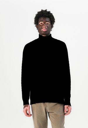 Maglione a collo alto nero con maniche lunghe, polsini a coste e vestibilità morbida, abbinato a pantaloni in corduroy beige. Tessuto semplice, ricco di texture.