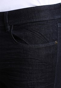 Vaqueros de mezclilla azul oscuro con un diseño clásico de cinco bolsillos, costuras prominentes y un pequeño botón metálico en el bolsillo delantero.