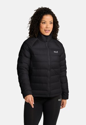 Vrouw met krullend haar die een zwarte donsjas en zwarte legging draagt, staand tegen een effen lichtgrijze achtergrond.