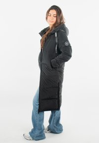 Manteau matelassé noir long avec capuche, comportant des poches zippées et un patch logo sur la manche. Porté par-dessus un jean bleu et des baskets.