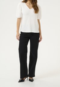 Weißes, strukturiertes Oberteil mit Puffärmeln, kombiniert mit hoch sitzenden, geraden schwarzen Jeans und spitzen, schwarzen Absatzschuhen. Schlichtes, modernes Design.