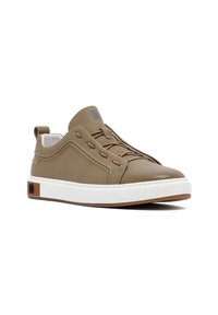 Zapatilla de cuero marrón con punta redondeada, diseño sin cordones, media suela blanca y suela de goma. Presenta detalles de costura y un bucle trasero.