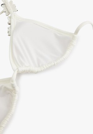 Haut de bikini blanc avec bords froncés et petites décorations en forme de fleurs le long de la couture supérieure sur fond uni.
