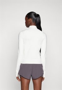 Hvit langermet treningstopp med høy hals, sammen med grå shorts. Stoffet virker glatt og fleksibelt, designet for aktiv bruk.
