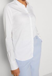 Camicia bianca a maniche lunghe con colletto, texture liscia e orlo dritto. Indossata con pantaloni azzurri chiari.