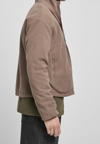 Giacca in pile marrone con colletto alto, tasca laterale con zip e design corto, abbinata a un maglione verde a maglia.