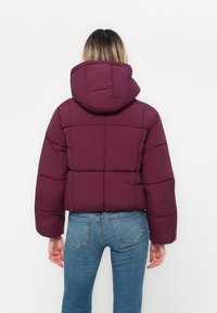 Bordo puffer jakna s kapuljačom, s prešanim dijelovima i skraćenim dizajnom, u kombinaciji s plavim trapericama.
