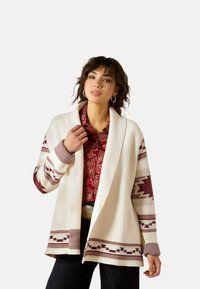 Cremfarbener Strickcardigan mit braunen geometrischen Mustern an den �Ärmeln und am Saum. Tiefer Kragen und gerippte Bündchen, kombiniert mit einer roten gemusterten Bluse darunter.