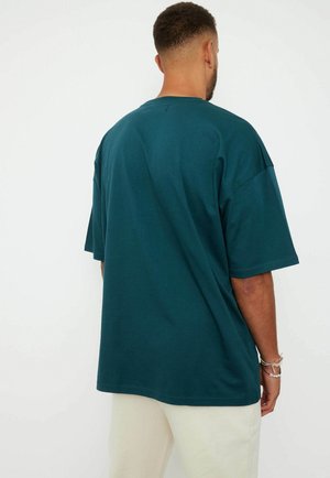 Man with short curly hair wearing a loose dark teal short-sleeve shirt and light-colored pants, shown from the back.