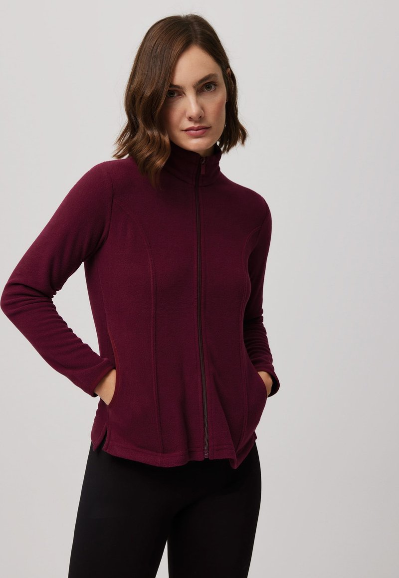 Chaqueta de forro polar burdeos con cuello alto, cremallera completa, bolsillos laterales y costuras contorneadas. Textura suave y diseño ajustado.