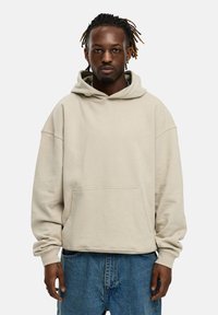 Beige Kapuzenpullover mit einer frontalen Tasche, lockerer Passform, gerippten Bündchen und einer Kordelzugkapuze, kombiniert mit blauen Jeans. Glatte Textur.