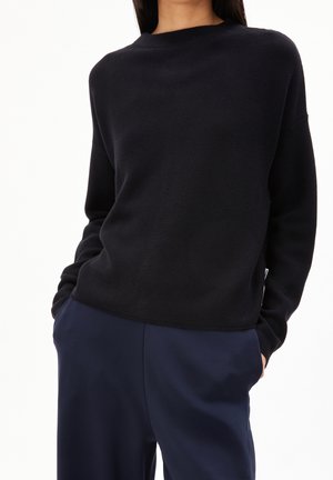 Femme portant un pull noir à manches longues et un pantalon bleu marine, les mains dans les poches, vue des épaules jusqu'au haut des cuisses.