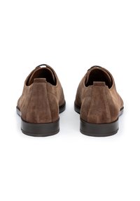 Chaussures slip-on en suede marron avec un bout arrondi, semelle extérieure noire basse et peu de détails. Vue de dos montrant une texture lisse et une couture discrète.